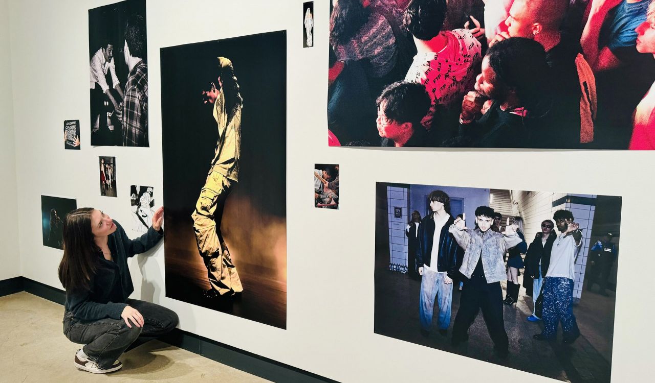 A woman crouches beside a wall of colourful photographs, looking up at a photo of a person at a music festival performing under stage light.