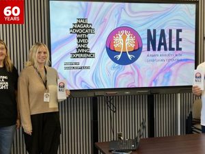 In a board room, Alicia Marshall, Keanna Schuster and Scott Neufeld with a large screen showing the NALE logo.