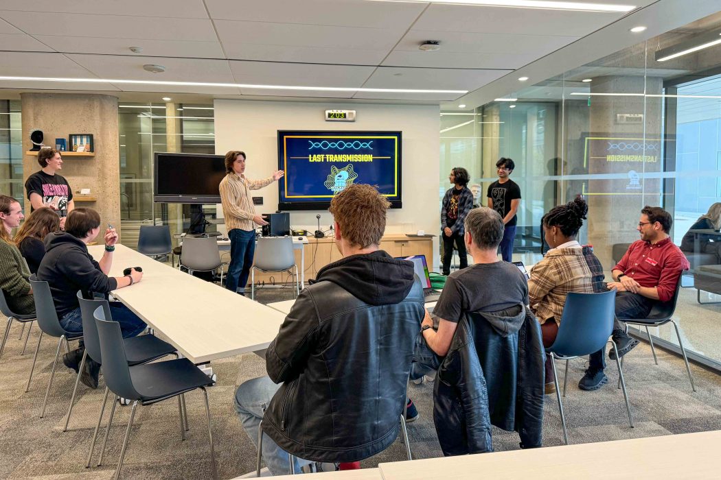 A group of students stand in front of a panel of people, presenting and referencing a large presentation screen that reads Last Transmission and a digital logo graphic.