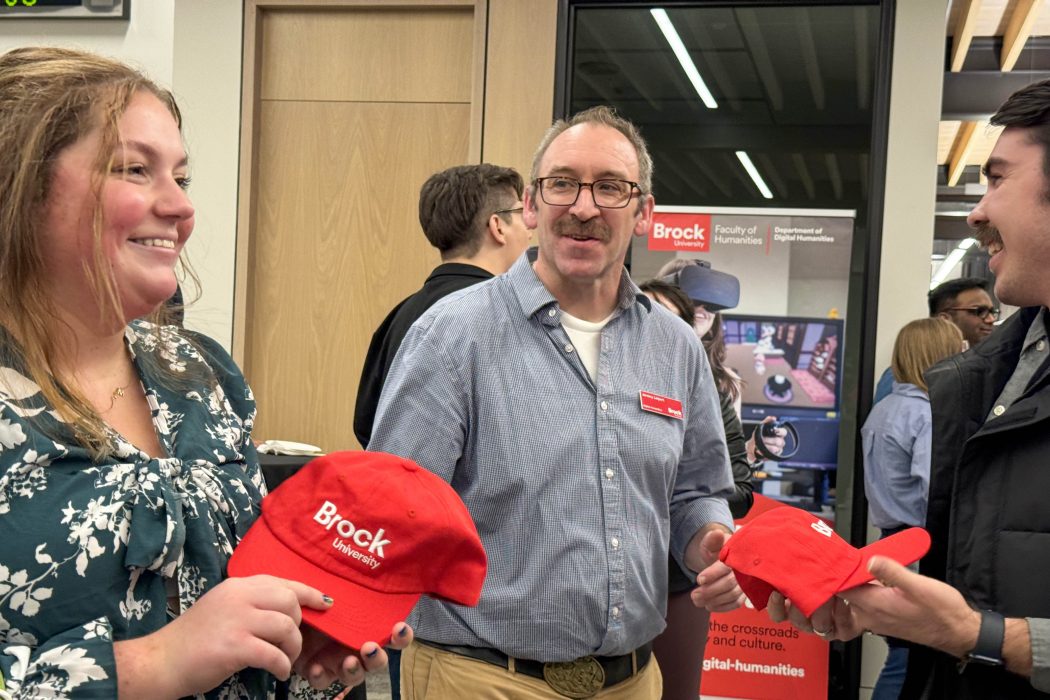 Three people stand together in a room smiling at each other, two of the people hold Brock hats that are bright red.