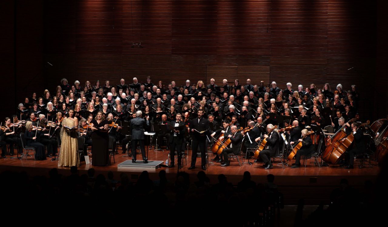 Musicians, choir members and soloists stand together in a large group under stage light performing Verdi’s Requiem. Three vocalists stand in the front of the group, a woman wearing a shining gold dress, while everyone else on stage wears black formal clothes.