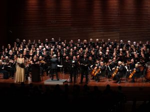 Musicians, choir members and soloists stand together in a large group under stage light performing Verdi’s Requiem. Three vocalists stand in the front of the group, a woman wearing a shining gold dress, while everyone else on stage wears black formal clothes.