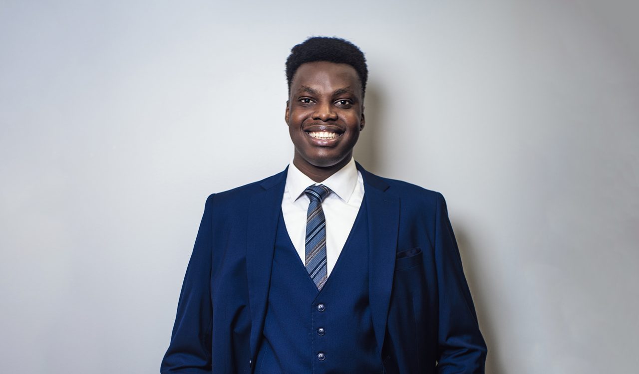 Tochukwu Daniel Oguejiofor stands against a neutral wall while wearing a blue formal suit.