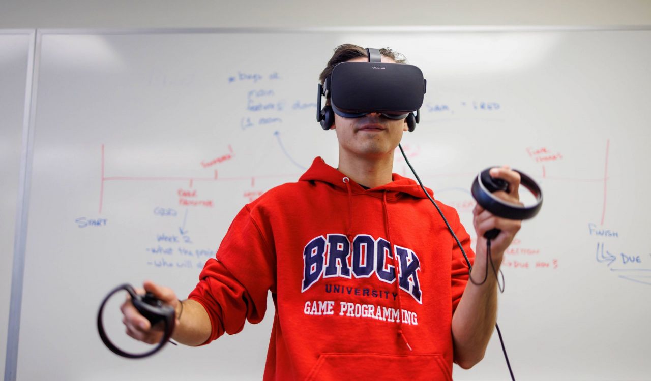 A university student wearing a red Brock sweatshirt stands in front of a whiteboard wearing a virtual reality head set with his arms outstretched.