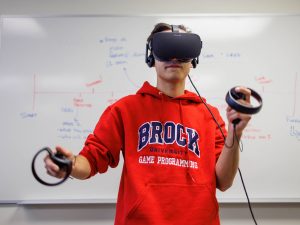 A university student wearing a red Brock sweatshirt stands in front of a whiteboard wearing a virtual reality head set with his arms outstretched.