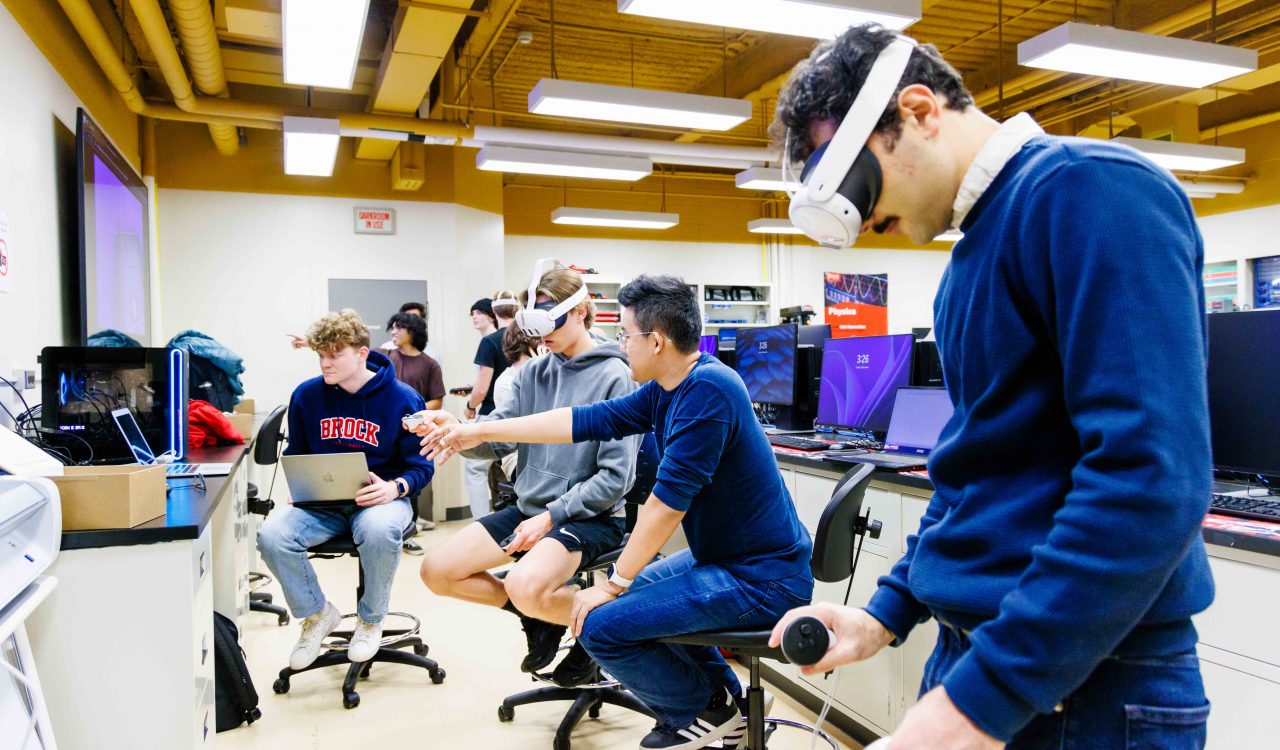 A student uses a virtual reality headset while other students in the background point at screens.