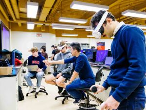 A student uses a virtual reality headset while other students in the background point at screens.