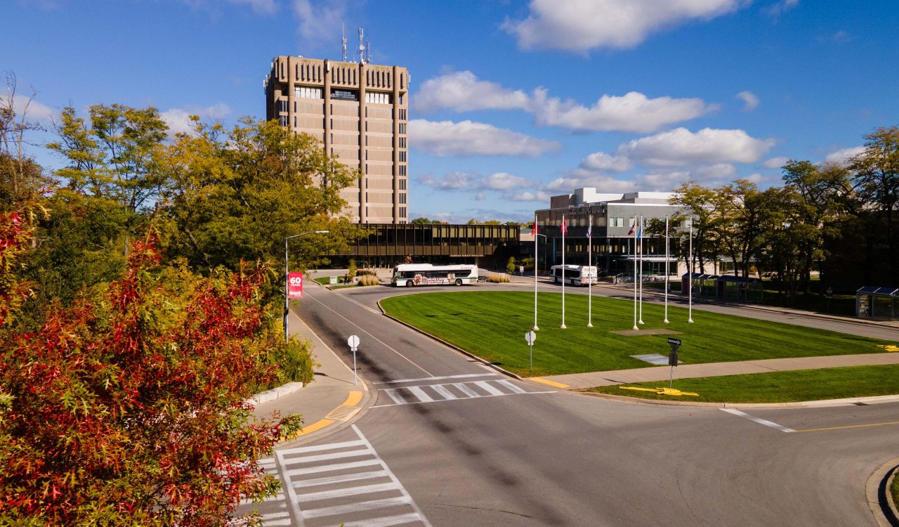 An aerial view of Brock University’s campus from the front with roadways, grass, trees and a blue sky with a tower building in the background.