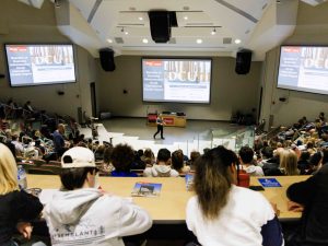 A wide-angle of a full theatre watching and listening to a man speak at the head of the room with three large screens brightly displaying a presentation about Brock University.