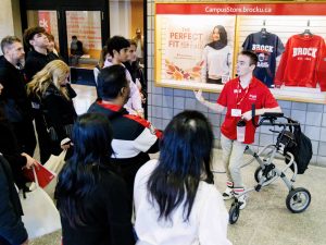 A man with an ambulation device leads a tour of a large crowd through a hallway at Brock University.