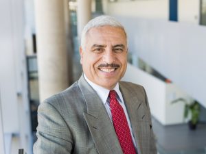 A man in a suit poses for a headshot in a sunny hallway.