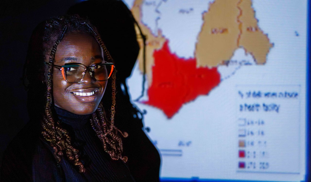 Portrait of Nana Adwoa Owusu, an International Master's student in Geography at Brock University, standing in front of a projected GIS map of Ghana. The map, which she uses for her research, highlights regional disparities as she explores predictors of early childhood mortality in her home country.