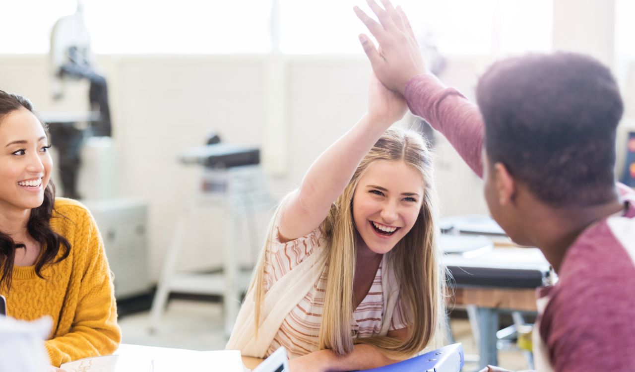 A young woman sits at a desk and high-fives another student, while a friend looks on smiling.