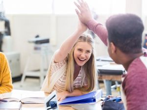 A young woman sits at a desk and high-fives another student, while a friend looks on smiling.