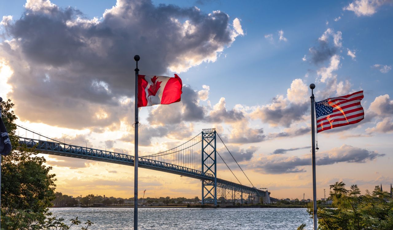 A Canadian and American flag fly in the foreground with the Ambassador Bridge stretching across the water between Windsor and Detroit