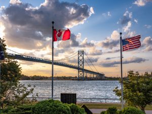 A Canadian and American flag fly in the foreground with the Ambassador Bridge stretching across the water between Windsor and Detroit