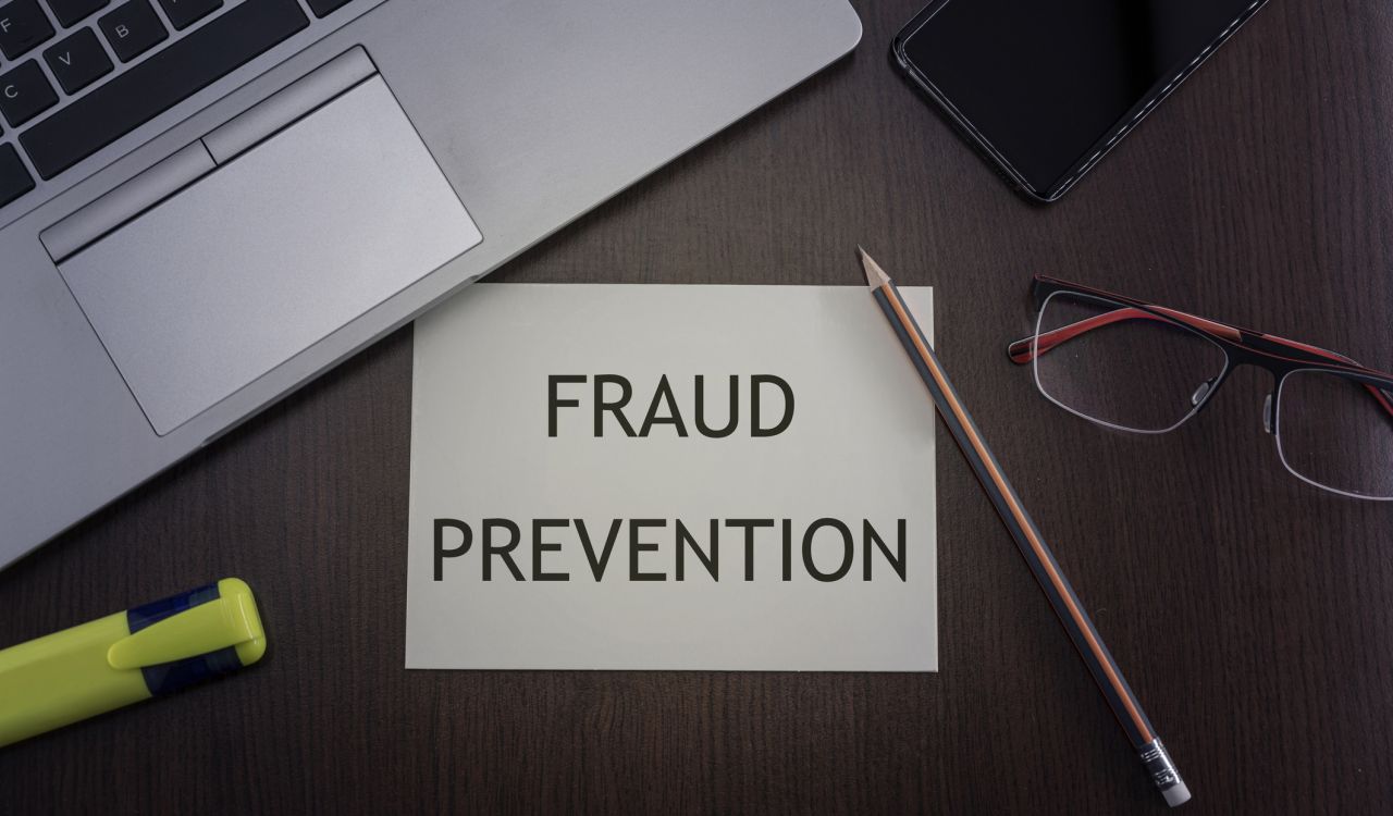 A card reading "fraud prevention" is on a desk beside a laptop, a pair of glasses, and office supplies.