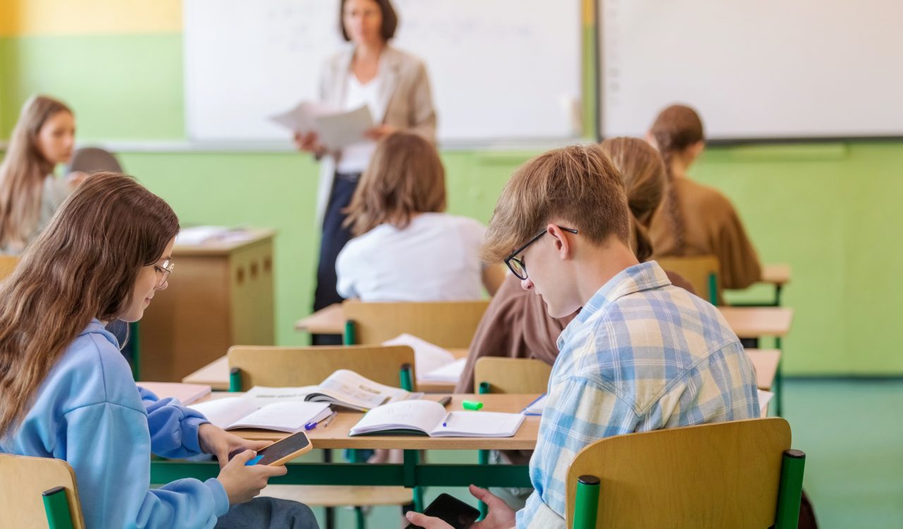 High school students sitting at the desk in the classroom and using smart phones with defocused teacher in the background.