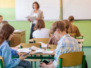 High school students sitting at the desk in the classroom and using smart phones with defocused teacher in the background.