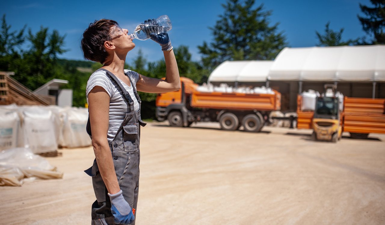 Woman wears overalls, work gloves safety glasses and drinks a bottle of water at a job site with large trucks.