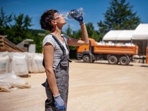 Woman wears overalls, work gloves safety glasses and drinks a bottle of water at a job site with large trucks.