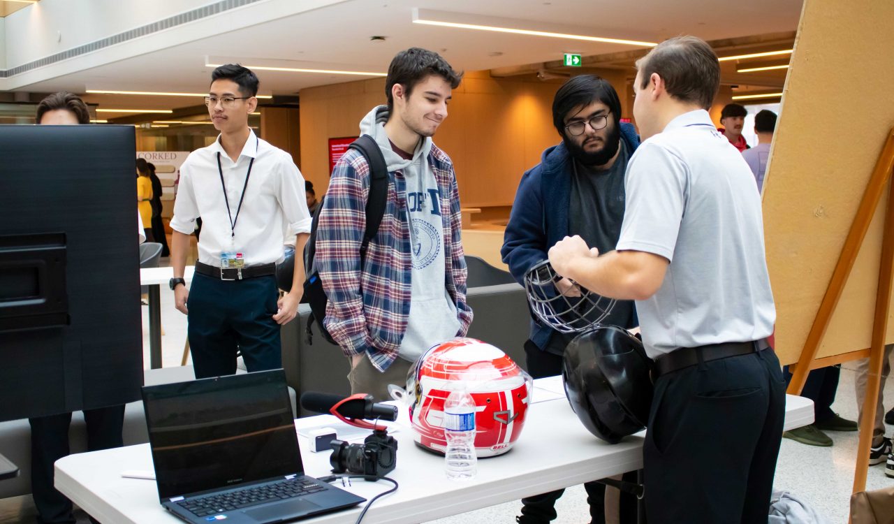 A university student shows a caged helmet to two individuals at a business class presentation.