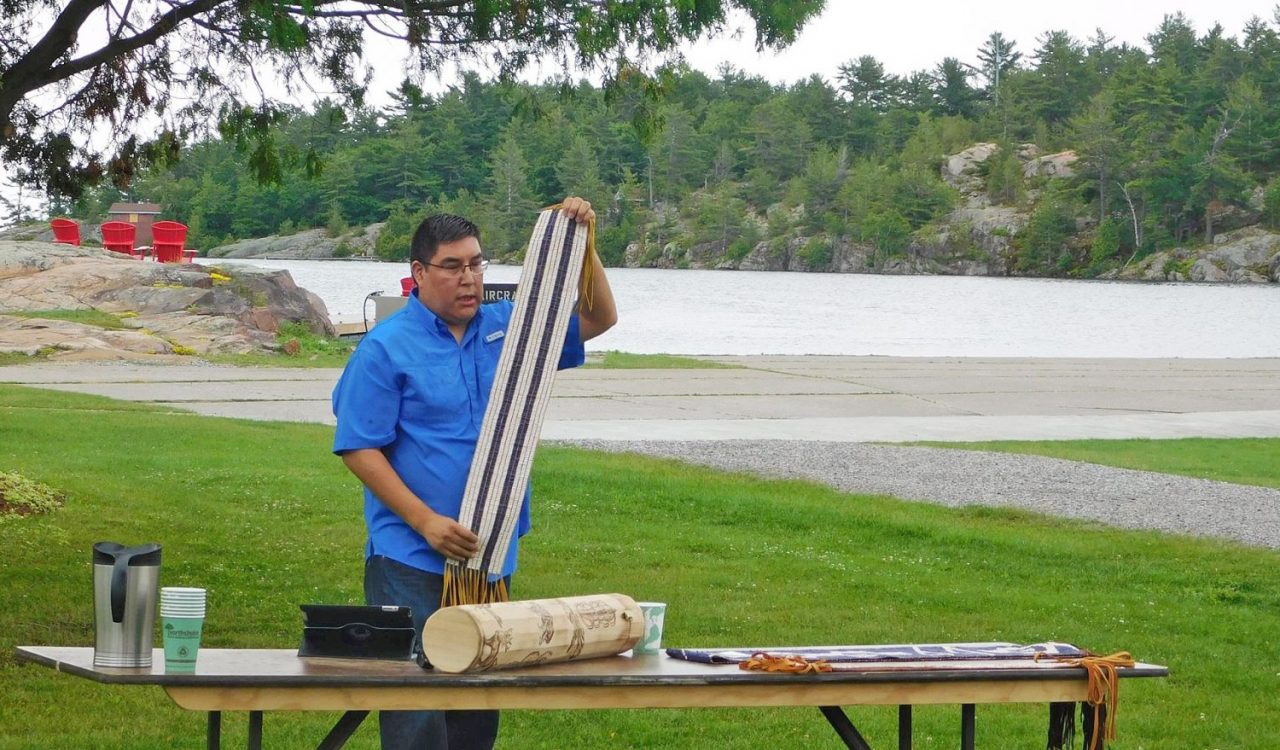 Joshua Manitowabi stands behind a table holding up a strip of beaded leather with three white and two purple stripes (a two-row Wampum belt), with another belt, a cylindrical object, an iPad, coffee cups and a coffee carafe placed on the table. Behind him is part of a lagoon, rocks and pine trees.