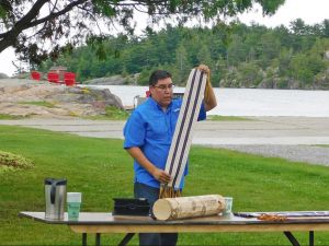 Joshua Manitowabi stands behind a table holding up a strip of beaded leather with three white and two purple stripes (a two-row Wampum belt), with another belt, a cylindrical object, an iPad, coffee cups and a coffee carafe placed on the table. Behind him is part of a lagoon, rocks and pine trees.