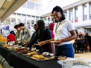 A woman smiles alongside other women at a food station in front of a large crowd inside a hall with high ceiling in a brightly-lit environment at Brock University.