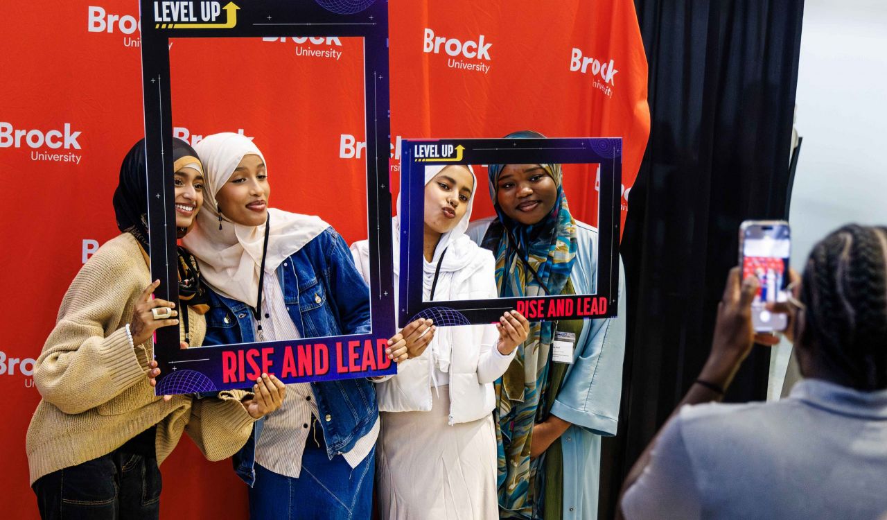 A group of four friends wearing cultural clothing smile against a red photo backdrop which reads Brock University while holding up frames with words such as ‘Level Up’ and ‘Rise and Lead’ on them as a photographer takes their photo.