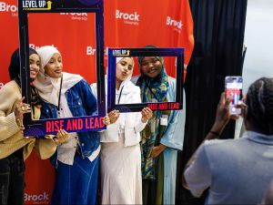 A group of four friends wearing cultural clothing smile against a red photo backdrop which reads Brock University while holding up frames with words such as ‘Level Up’ and ‘Rise and Lead’ on them as a photographer takes their photo.