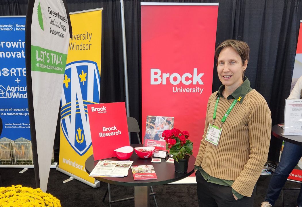 A smiling Malkie Spodek stands in front of a round table with pamphlets. In the background are pop-up banners from Brock and other institutions.