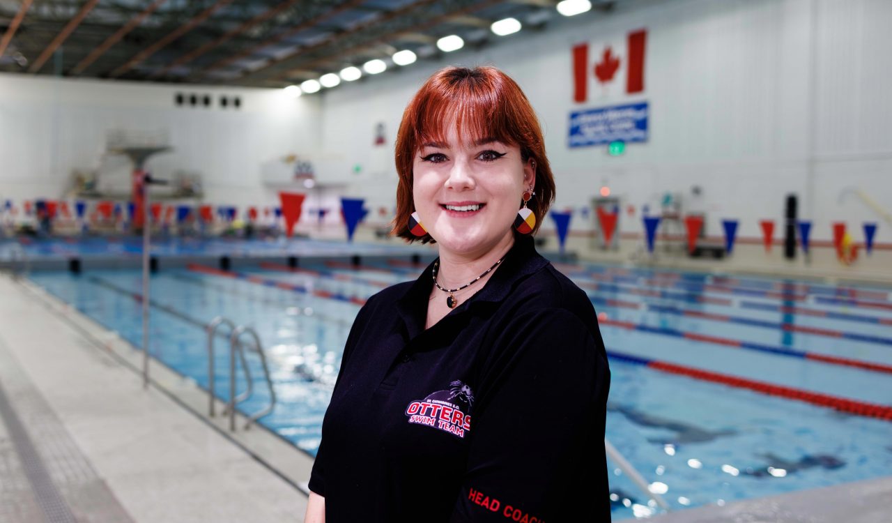 A woman with red hair and a black polo with the words ‘head coach’ and ‘St. Catharines Otters’ written on it stands in front of a pool at Brock University.