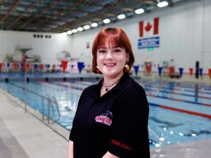 A woman with red hair and a black polo with the words ‘head coach’ and ‘St. Catharines Otters’ written on it stands in front of a pool at Brock University.