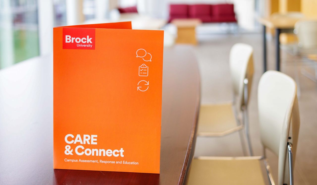 An orange folder with the words ‘Brock University’ and ‘CARE & Connect (Campus Assessment, Response and Education), sits standing up on a desk in a brightly lit room with windows, tables, chairs and a desk.