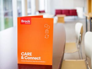 An orange folder with the words ‘Brock University’ and ‘CARE & Connect (Campus Assessment, Response and Education), sits standing up on a desk in a brightly lit room with windows, tables, chairs and a desk.