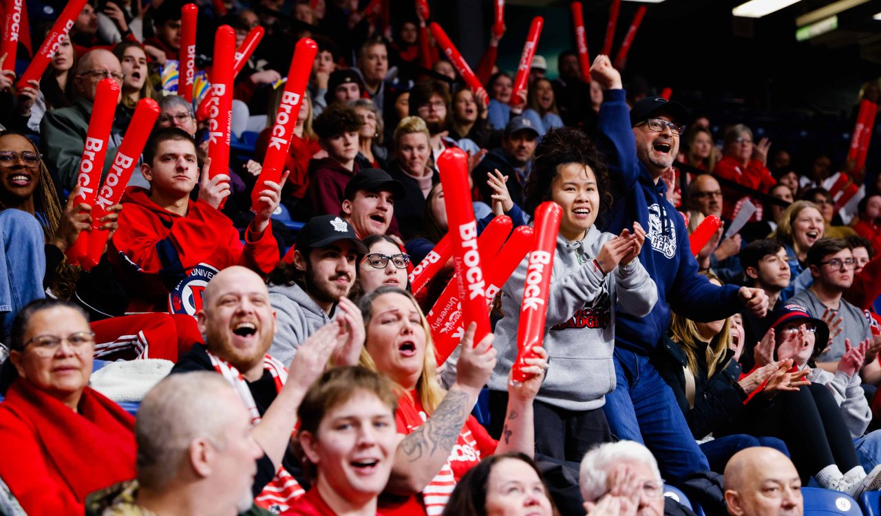 Brock University fans fill the stands at the Meridian Centre, cheering and waving red Brock-branded inflatable noise sticks during a women's basketball game. The crowd is full of energy, with supporters of all ages wearing Brock gear, clapping and smiling as they show their school spirit.