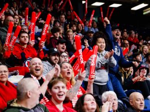 Brock University fans fill the stands at the Meridian Centre, cheering and waving red Brock-branded inflatable noise sticks during a women's basketball game. The crowd is full of energy, with supporters of all ages wearing Brock gear, clapping and smiling as they show their school spirit.