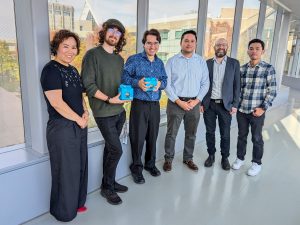 Six people pose for a photo in a sunny hallway. Two are holding blue boxes.