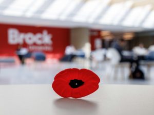 A red poppy pin sits on a white table in a brightly-lit room with a red wall in the background with the words Brock University in white.