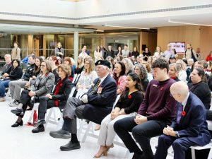 A crowd of people, many wearing red poppies, sit together in an indoor space during a Remembrance Day ceremony.