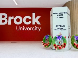 Three green wreaths with red poppies sit at the foot of a grey indoor cenotaph beside a red wall with the words 