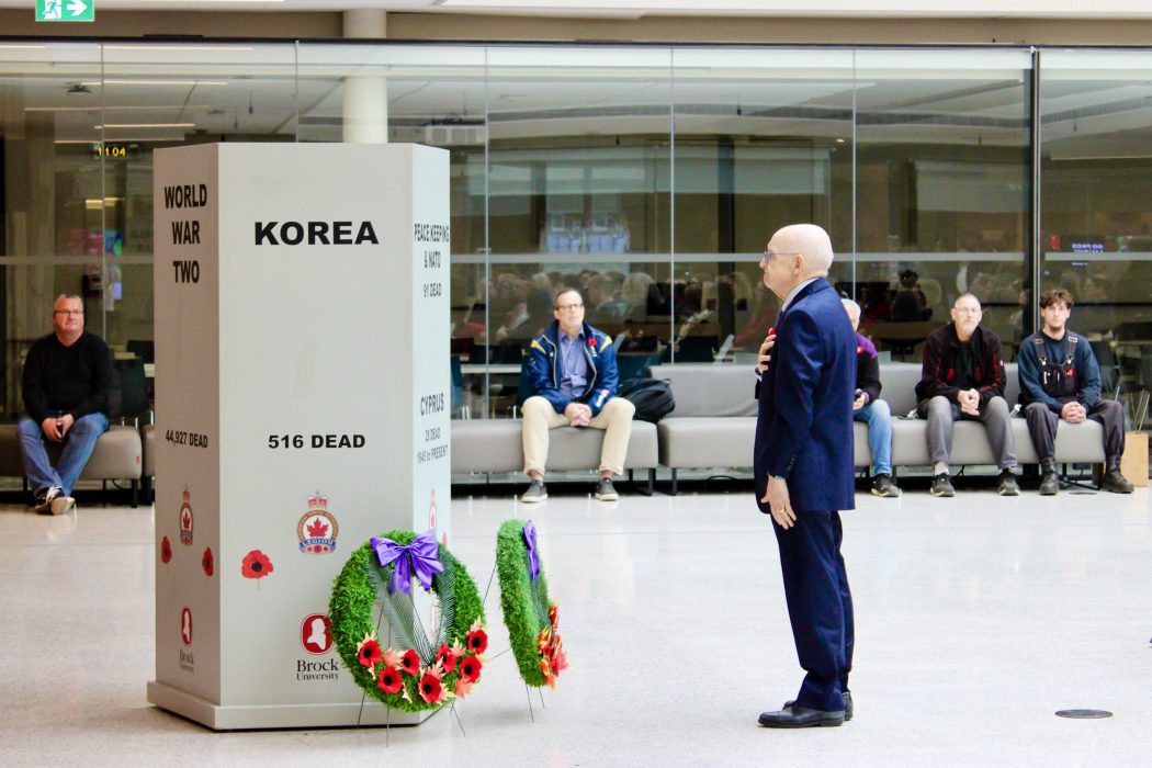 A man stands in front of a Remembrance Day memorial during a ceremony in a bright room.