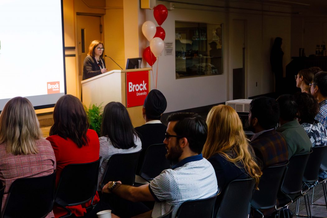 Long distance view of Brock University President and Vice-Chancellor Lesley Rigg standing at the podium, with part of a slide next to her, her hair a golden colour, with a group of people sitting in the foreground facing her.