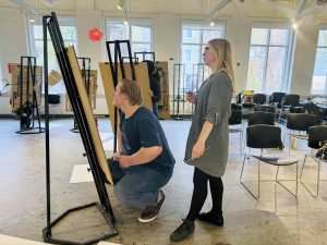 Two people stand at an easel in an art studio reviewing a piece of art.