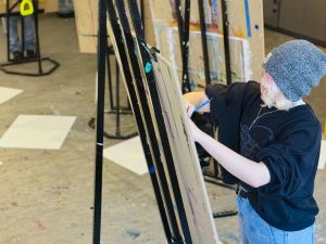 A person stands in front of an easel with their face tilted down in concentration as they draw.