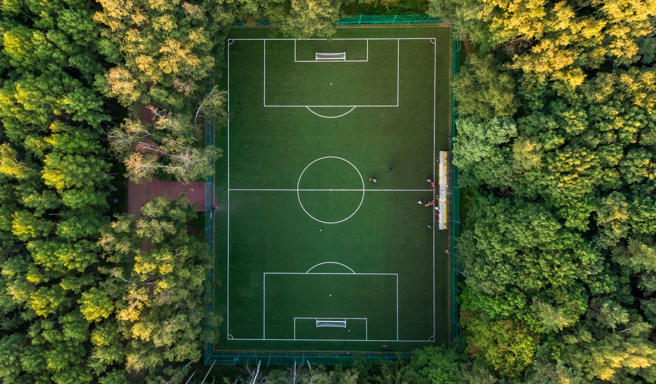 Aerial shot taken with a drone of an outdoor soccer pitch surrounded by trees with a few players on the field.