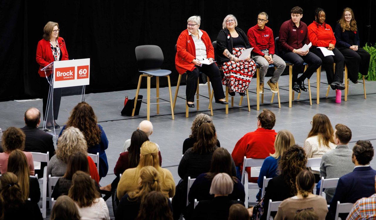 Audience seated in a university gymnasium listens attentively to a speaker at a podium and a panel of presenters at the front of the room, all wearing red attire associated with Brock University.