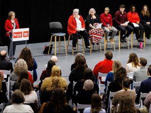 Audience seated in a university gymnasium listens attentively to a speaker at a podium and a panel of presenters at the front of the room, all wearing red attire associated with Brock University.