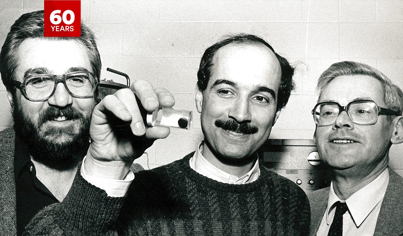 Black-and-white photo of Bozidar Mitrovic, Fereidoon Razavi and the late Frans Koffyberg in which Razavi (centre) is holding up an object containing a circular blob; the other two men are looking at the object and smiling.